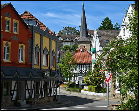 Dorfstraße mit bunten Fachwerkhäusern in Gelb, Rot und Schwarz-Weiß; im Hintergrund ein spitzer Kirchturm und ein Fachwerkgebäude mit orangenem Dach.