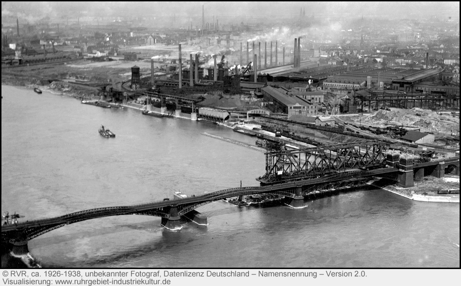 Ein altes Foto einer Brücke über einen Fluss. Im Hintergrund viel Industrie mit Schornsteinen und Qualm.