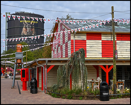 Ein Holzgebäude mit rot-weißen Kachelmustern vor einem großen Gasometer. Kleine Wimpel mit Erdbeeren sind über den Weg gespannt.