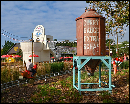 Einschienenbahn mit einer Currywurst als Fahrzeug, daneben ein Silo mit "Currysoße Extrascharf" und eine große Toilette als Gebäude