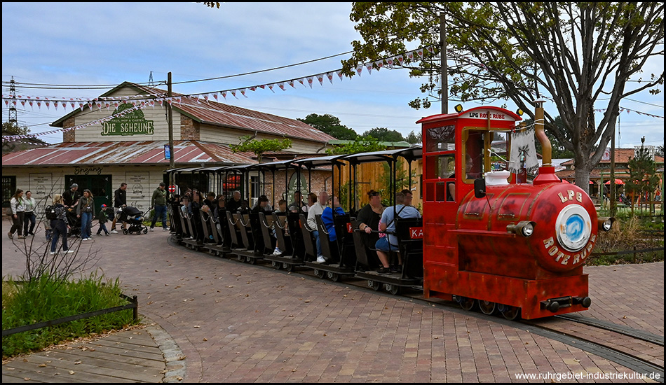 Park-Eisenbahn mit einer roten Lok, die wie eine Dampflok aussieht und die Beschriftung "LPG Rote Rübe" trägt. Dahinter mehrere schwarze Waggons mit Fahrgästen. Der Zug fährt vor einem Gebäude mit der Aufschrift "Die Scheune" über einen gepflasterten Platz.
