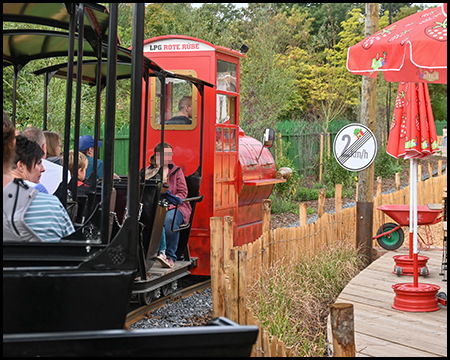Ein kleiner Freizeitpark-Zug mit roter Lok fährt um eine Kurve. Rechts ist eine Terrasse mit roten Sonnenschirmen zu sehen. Ein Schild weist darauf hin, dass eine Geschwindigkeitsbegrenzung von 2 km/h aufgehoben ist.