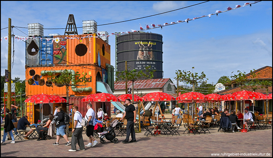 Szene aus einem Freizeitpark mit Sonnenschirmen und Tischen und Stühlen, ein Spielplatz aus Fracht-Containern und im Hintergrund der Gasometer Oberhausen