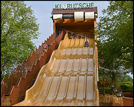 Eine große Rutsche mit sechs Rutschbahnen nebeneinander, oben übergroße Rollen von Toilettenpapier, darüber die Schrift "Klorutsche"