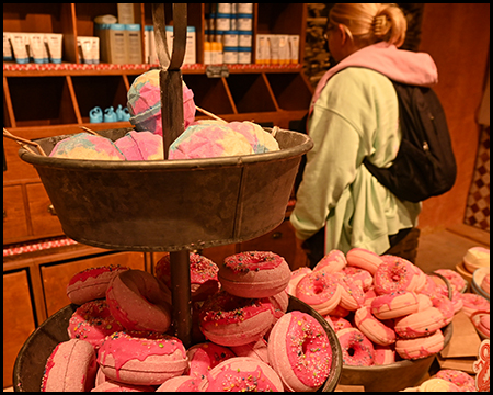 Eine Frau steht in einem Verkaufsbereich und guckt sich die Auslage an. Im Vordergrund pinkfarbene Badezusätze in Donutform