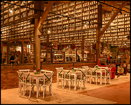 Blick in eine Halle aus Holz. Im Vordergrund stehen Tische und Stühle. Im oberen Teil des Tragwerks stehen Regale mit einer Sammlung von Kaffeekannen.