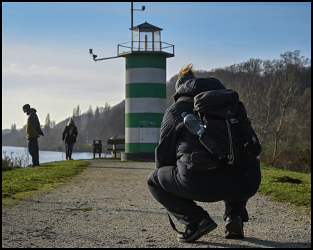 Eine Frau kniet auf einem Weg und fotografiert einen kleinen grün-weißen Leuchtturm an einem See