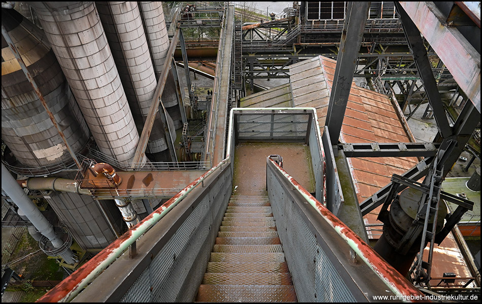 Blick vom oberen Ende einer Treppe abwärts. Die Treppe führt mitten durch eine Industrieanlage eines Hochofens.