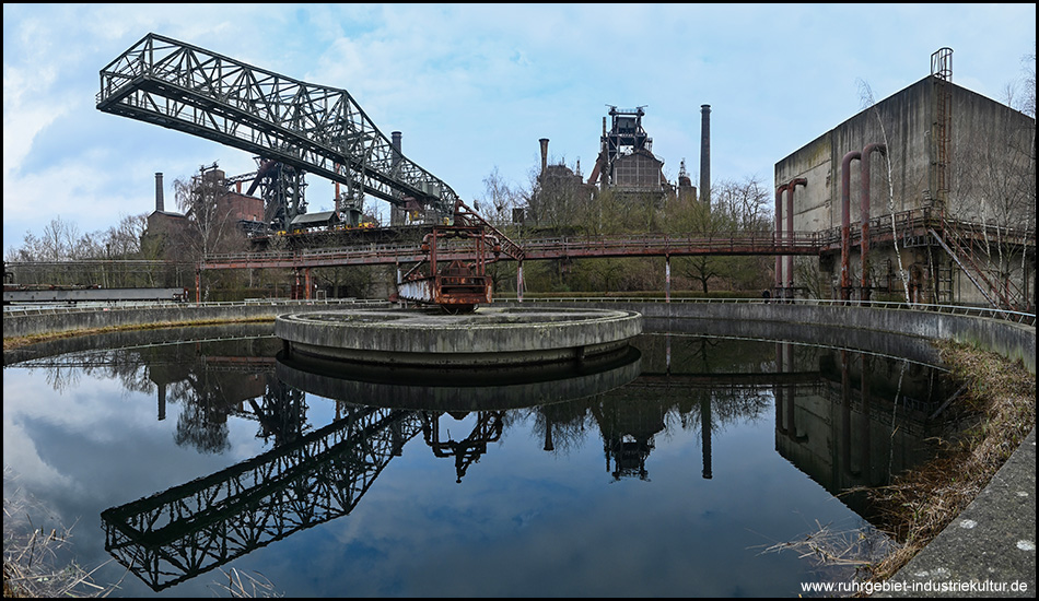 Panoramabild mit einem Klärbecken und den darin spiegelnden Anlagen des Hochoofenwerks, unter anderem mit dem Krokodil