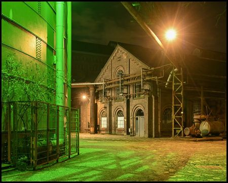 Grün beleuchteter Stahltank und eine Industriehalle mit Straßenbeleuchtung in der Nacht
