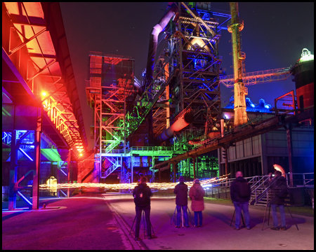Fotografen stehen mit Stativen und Kameras inmitten einer farbig illuminierten Industriekulisse