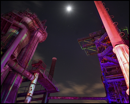 Blick nach oben auf hoch aufragende Industrieanlagen bei Nacht, die farbig beleuchtet werden. Es sind Mond und Sterne sichtbar