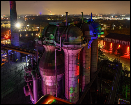 Silos und Anlagen des Hüttenwerks Meiderich vom Hochofen aus gesehen in der Dunkelheit. Sie sind farbig beleuchtet. Die Silos wirken lila und grün, eine Halle hat rote regelmäßige Scheinwerfer an der Wand.