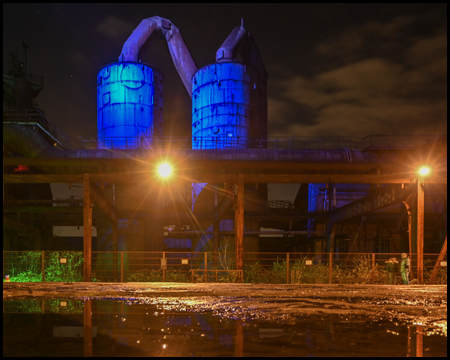 Ein blau beleuchtetes Silo hinter einem Platz mit Pfützen, in denen sich die Szene teilweise spiegelt