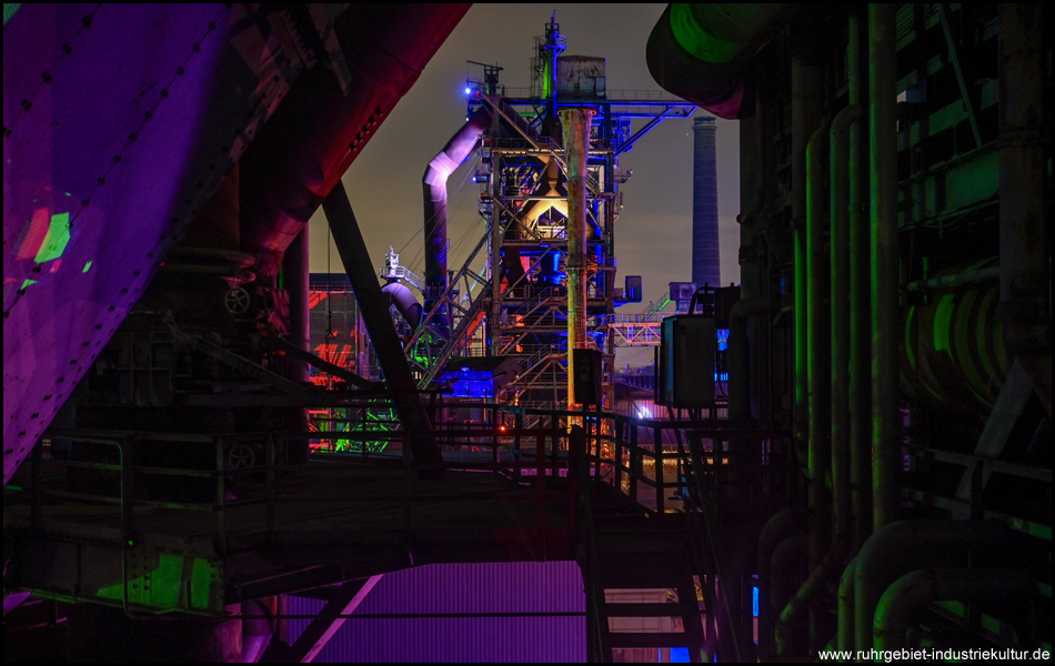 Inmitten einer nächtlichen Industrieanlage mit teilweise beleuchteten Anlagenteilen. Blick durch eine Lücke auf einen farbig illuminierten Hochofen