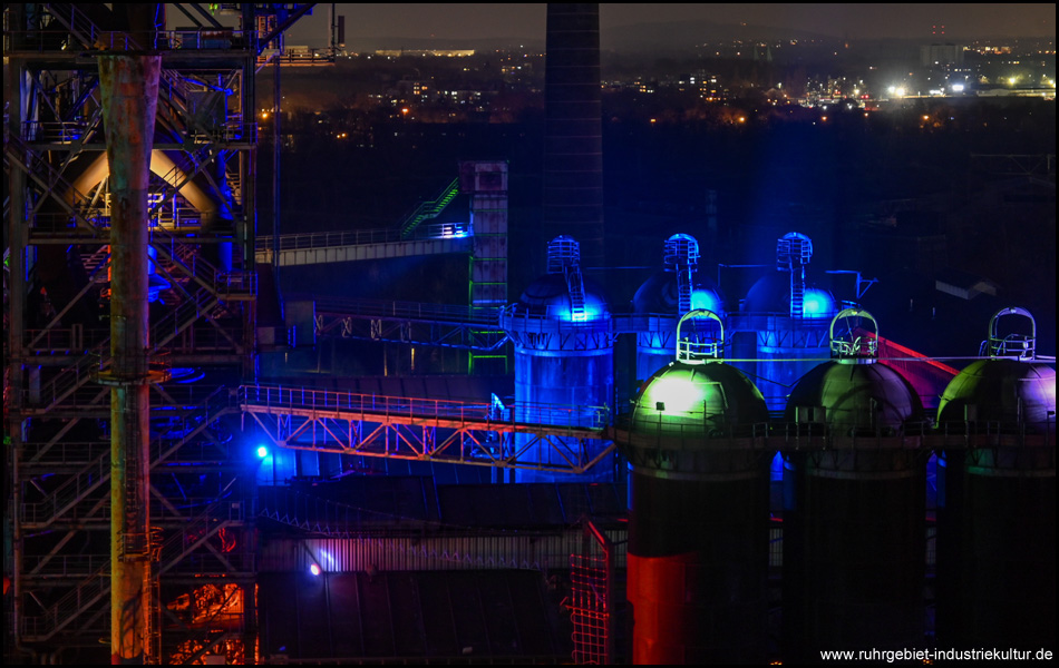 Aussicht vom Hochofen 5 des Hüttenwerks auf Silos und andere Teile von Hochöfen in der Dunkelheit. Die Silos sind blau und grün beleuchtet.