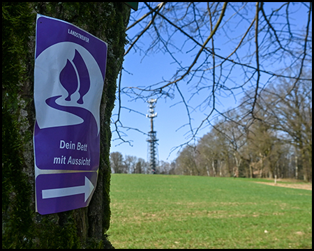 Ein lila Wegweiserschild mit Symbol "Dein Bett mit Aussicht" an einem Baum befestigt. Im Hintergrund eine Wiese und ein Funkturm.
