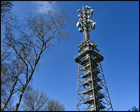 Ein hoher Sendemast mit zahlreichen Antennen und Satellitenempfängern vor einem klaren blauen Himmel und kahlen Bäumen.