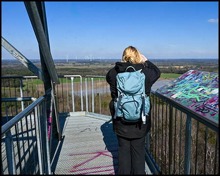 Eine Person mit Rucksack auf einer Aussichtsplattform, die mit einem Fernglas oder einer Kamera in die Landschaft blickt.