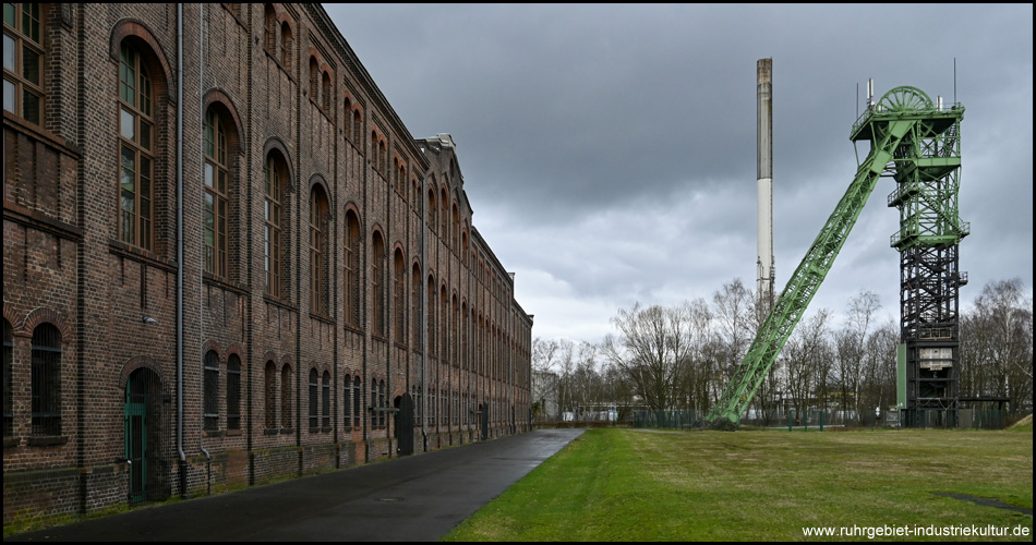 Große Backstein-Halle mit hohen Fenstern. Daneben ein Fördergerüst hinter einer Wiese