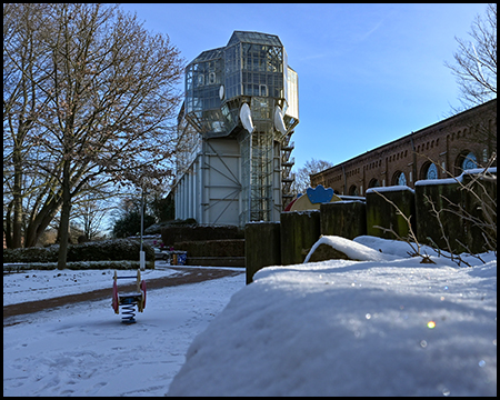 Bauwerk aus Beton mit einem Anbau, der dem Gebäude die Form eines Elefanten gibt, auf verschneitem Platz, flankiert von Backsteingebäuden unter klarem Winterhimmel.