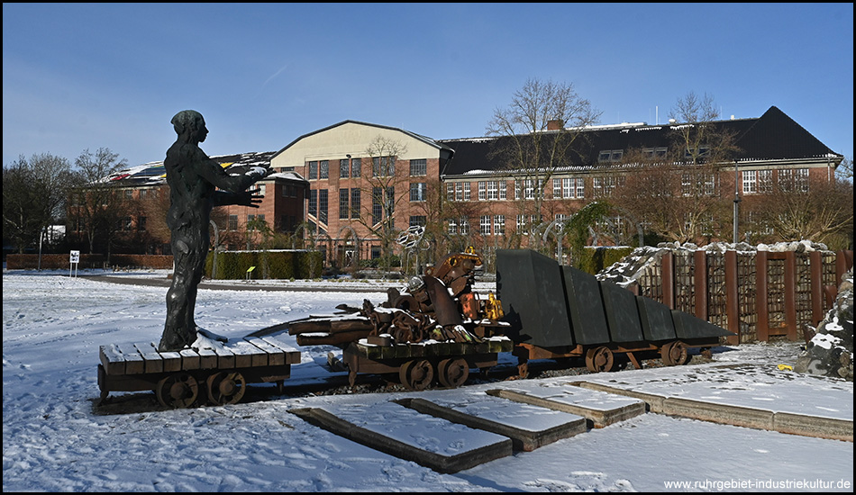 Metallskulptur einer Bergarbeiterfigur mit Grubenwagen auf Schienen vor Backsteingebäuden im verschneiten Industriepark.