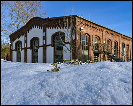 Historisches Backsteingebäude mit weißer Fassade, großen Rundbogenfenstern und dekorativem Zinnenkranz unter Schneedecke.