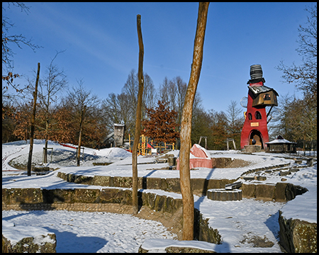 Verschneiter Spielplatz mit Terrassen, Holzpfosten und rotem Spielturm im Hintergrund.