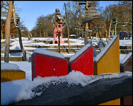 Detail eines bunten Holzspielgeräts in Rot, Gelb und Schwarz mit Schnee bedeckt, Spielturm unscharf im Hintergrund.