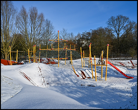 Spielplatzbereich mit orange-gelben Klettergerüsten und roten Rutschen, vollständig schneebedeckt unter blauem Himmel.