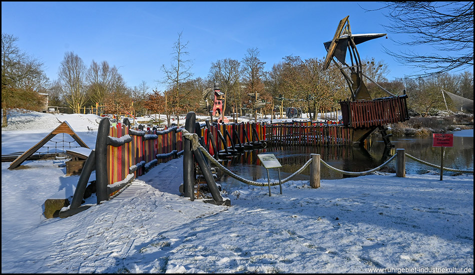 Bunter Spielplatz im Schnee mit Holzsteg über einen gefrorenen See, im Hintergrund kahle Bäume unter blauem Himmel.