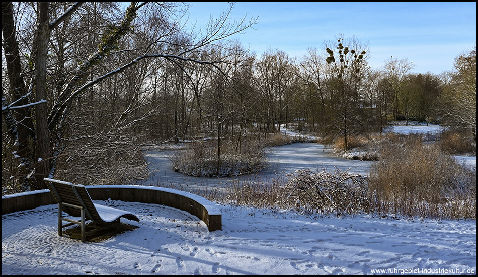 Verschneite Parkbank am Ufer eines zugefrorenen Teichs mit kleinen Inseln, umgeben von kahlen Bäumen unter teilweise bewölktem Himmel.