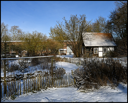 Fachwerk-Bauernhaus mit schneebedecktem Dach hinter Holzzaun und vertrockneten Pflanzen unter blauem Himmel.