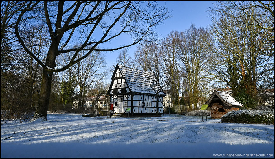Historisches Fachwerkhaus mit blau-weißer Fassade auf verschneiter Wiese, umgeben von kahlen Bäumen unter blauem Winterhimmel mit langen Schatten.