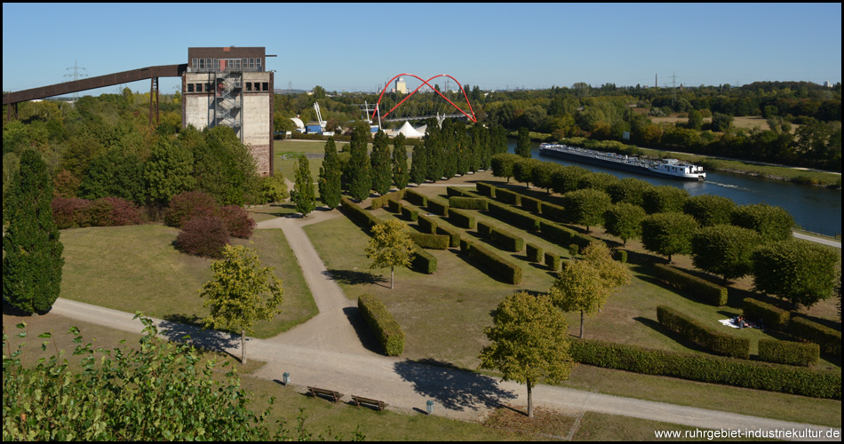 Blick von einem Ausläufer der Ruhrgashalde auf den Kohlebunker, die Doppelbogenbrücke und den Rhein-Herne-Kanal. Die Hecken symbolisieren ein Gleisbild mit Schienen und Weichen, die eckigen Bäume die Güterzüge