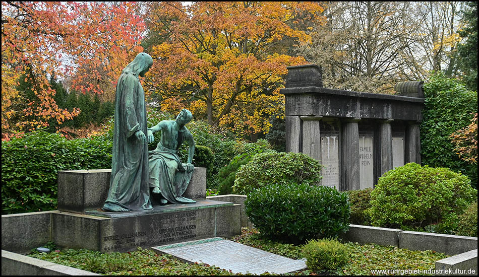 Grabanlage mit zwei bronzegrünen Frauenfiguren auf steinerner Plattform, eine stehend, eine sitzend. Rechts davon ein dunkles Mausoleum im Bauhausstil. Herbstlich gefärbte Bäume im Hintergrund, gepflegte Büsche im Vordergrund.