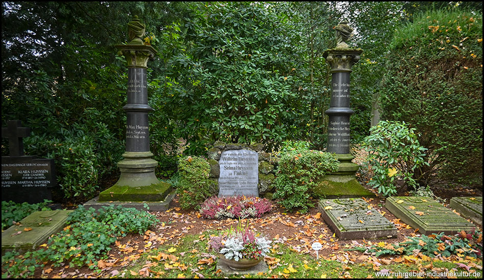 Familiengrabstätte mit weißem zentralem Grabstein, flankiert von zwei dunkelgrauen Säulen mit Urnen. Herbstlaub bedeckt den Boden, geschmückt mit Heidekraut und weißen Stiefmütterchen. Dichte Rhododendren im Hintergrund.