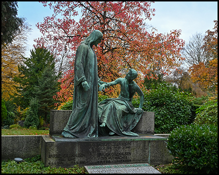 Bronzegrüne Skulpturengruppe zweier trauernder Frauen auf Steinsockel. Eine steht, die andere sitzt in Trauerpose. Herbstlich gefärbte Bäume mit rot-orangem Laub im Hintergrund.