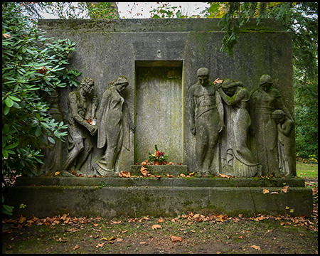 Verwittertes Wandgrabmal mit vier Trauerfiguren in dramatischen Posen vor einer steinernen Wand mit zentraler Inschrifttafel. Herbstlaub liegt auf dem Sockel, umgeben von Rhododendren.