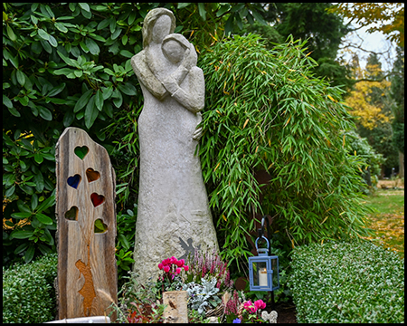 Moderne, schlichte weiße Steinskulptur einer Mutter mit Kind. Daneben ein Holzstele mit herzförmigen Ausschnitten. Frische Blumen und ein Grablicht am Fuß. Grüne Hecken im Hintergrund.