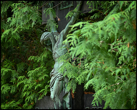 Bronzegrüne Engelsfigur mit ausgebreiteten Flügeln, teilweise verdeckt durch üppige grüne Nadelbaumzweige