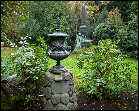 Schmiedeeiserne Urne auf verwittertem Steinsockel, umgeben von Rhododendren und anderen immergrünen Pflanzen. Im Hintergrund weitere Grabdenkmäler in grüner Parklandschaft.