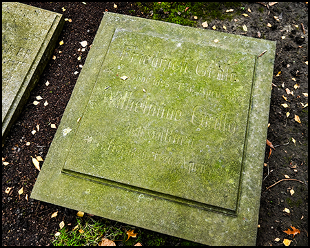 Nahaufnahme einer moosbedeckten, quadratischen Grabsteinplatte mit kaum noch lesbarer Inschrift. Die verwitterte Oberfläche zeigt grüne Patina und Spuren der Zeit.