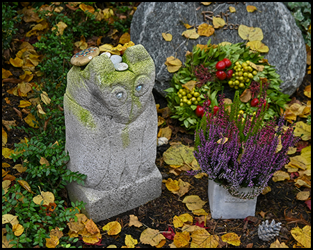 Verwitterte Steinskulptur einer Eule mit großen runden Augen, bedeckt mit Moos und Herbstlaub. Daneben violettes Heidekraut, rote Beeren und herbstliche Blätter auf dem Boden.