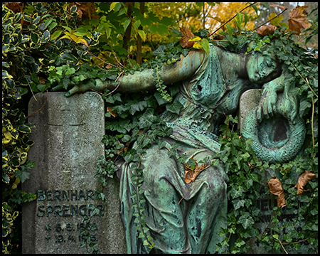 Bronzegrüne Reliefdarstellung einer trauernden Figur in fließenden Gewändern an einer Grabwand, teilweise von Efeu überwuchert. Herbstlaub liegt auf den Figuren. Links ein verwitterter Grabstein.
