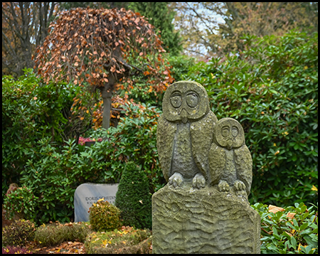 Steinskulptur zweier stilisierter Eulen mit großen runden Augen auf einem gemeinsamen Sockel. Eine größere und eine kleinere Eule. Herbstliche Bepflanzung und Hecken im Hintergrund.