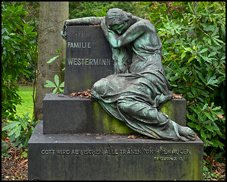 Bronzegrüne Trauerfigur einer Frau, die sich an einen dunklen Grabstein mit der Aufschrift "Familie Westermann" lehnt. Darunter ein Bibelzitat. Die Skulptur ist von grünen Rhododendren umgeben.