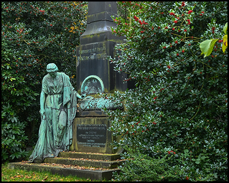 Bronzegrüne Trauerfigur einer Frau mit gesenktem Kopf vor einem dunklen Obelisken-Grabmal. Ein Kranz liegt auf dem Sockel. Immergrüne Sträucher mit roten Beeren im Hintergrund.