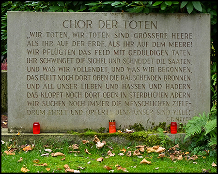 Steinerne Gedenktafel mit der Aufschrift "Chor der Toten" und einem mehrzeiligen Gedicht. Drei rote Grablichter stehen vor der Tafel, Herbstlaub liegt auf dem grünen Rasen davor.