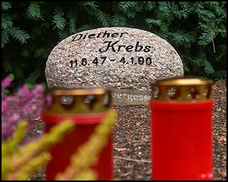 Moderner runder Grabstein mit der Aufschrift "Diether Krebs 11.8.47 - 4.1.00". Im Vordergrund unscharf ein rotes Grablicht und gelbe Herbstblätter. Grüne Nadelbäume im Hintergrund.